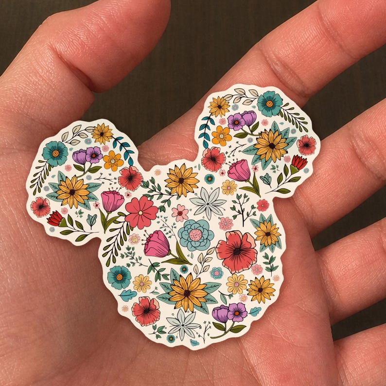 May include: A floral sticker in the shape of a mouse head. The sticker is filled with colorful flowers in shades of pink, yellow, blue, and purple, with green leaves. The sticker is held in a hand.