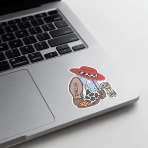 Puede incluir: Una pegatina que muestra un sombrero de vaquero rojo, una bota de vaquero con estampado de vaca, un patr&oacute;n de cielo azul con nubes y el nombre TAYLOR escrito en la suela. Junto a la bota hay tres bloques apilados con las letras T, S y un peque&ntilde;o dise&ntilde;o gr&aacute;fico.