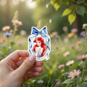 Peut inclure: Un autocollant repr&eacute;sentant une illustration &agrave; l'aquarelle d'un personnage aux longs cheveux roux et une robe bleue, encadr&eacute;e d'un n&oelig;ud et de rubans bleus. L'autocollant est tenu dans une main sur un fond flou de fleurs et de verdure.