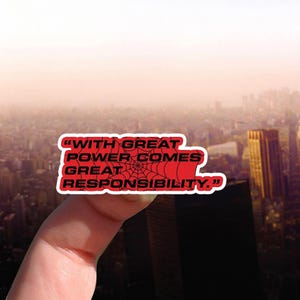 May include: A red sticker with the quote "WITH GREAT POWER COMES GREAT RESPONSIBILITY" in white text. A black spiderweb design is in the center. The sticker is held up against a blurred cityscape background.