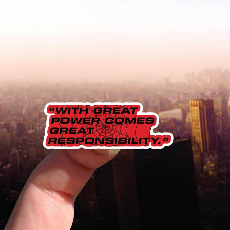 May include: A red sticker with the quote "WITH GREAT POWER COMES GREAT RESPONSIBILITY" in white text. A black spiderweb design is in the center. The sticker is held up against a blurred cityscape background.