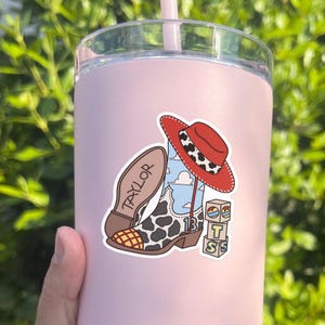 Puede incluir: Un vaso rosa claro con una pegatina que muestra una bota de vaquero con estampado de vaca, un sombrero rojo con banda de manchas, el nombre TAYLOR en la suela, el n&uacute;mero 13 y una pila de bloques con las letras T, S y S.