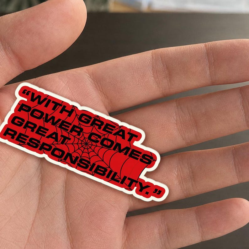 May include: A red sticker with the quote "WITH GREAT POWER COMES GREAT RESPONSIBILITY" in black text. A spiderweb design is in the center of the sticker. The sticker is held in a hand.