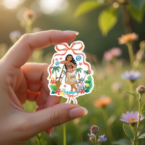May include: A colorful sticker featuring a cartoon illustration of a woman with long black hair, wearing a traditional outfit, and holding a paddle. The sticker includes palm trees, flowers, and a pig character. The sticker is held in a hand against a blurred background of flowers.