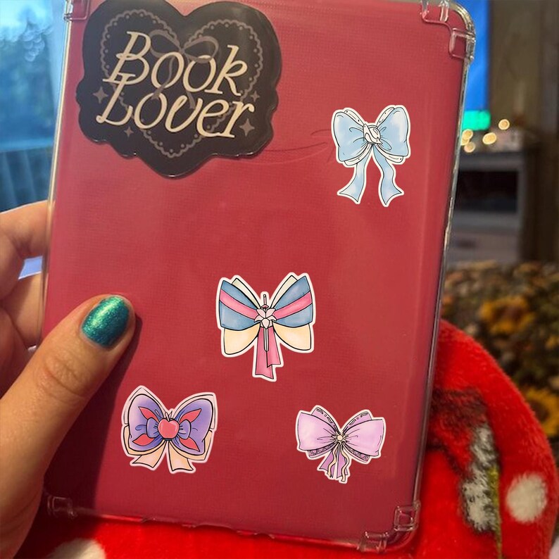 May include: A red tablet case with a clear border, adorned with bow stickers and a "Book Lover" sticker. The bow stickers are in various colors, including blue, pink, and purple. The "Book Lover" sticker is black with white text.