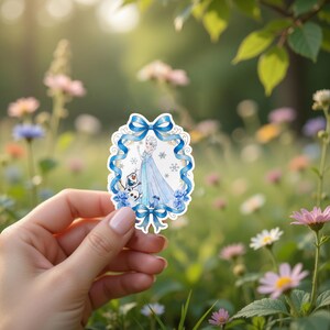 以下が含まれることがあります： アナと雪の女王のエルサと雪だるまのオラフの水彩イラストが描かれたステッカー。青いリボンと雪の結晶で縁取られ、ぼやけた花の背景を背景に、手で持たれています。