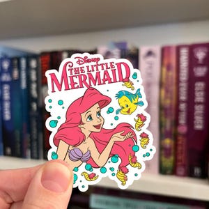 Op de afbeelding: Een gestanste sticker met Ariel uit De Kleine Zeemeermin met lang roze haar, een paarse schelpentop en een lichte huidskleur. Het ontwerp bevat de tekst Disney The Little Mermaid, een gele Botje-vis en verschillende kleine gele zeepaardjes omringd door blauwgroene bubbels op een witte achtergrond.