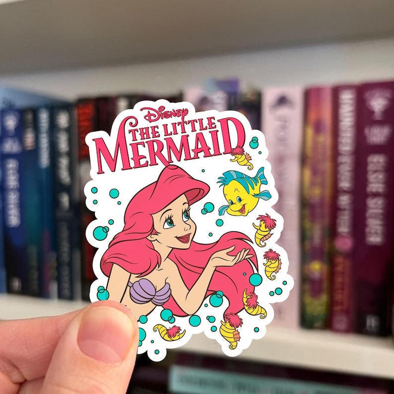 Op de afbeelding: Een gestanste sticker met Ariel uit De Kleine Zeemeermin met lang roze haar, een paarse schelpentop en een lichte huidskleur. Het ontwerp bevat de tekst Disney The Little Mermaid, een gele Botje-vis en verschillende kleine gele zeepaardjes omringd door blauwgroene bubbels op een witte achtergrond.
