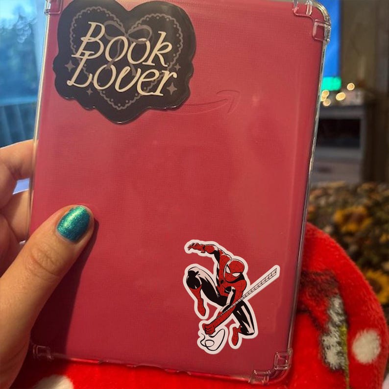 May include: A pink e-reader case with a clear protective cover. The case has a "Book Lover" sticker and a Spider-Man sticker. The e-reader is held by a person with blue nail polish.