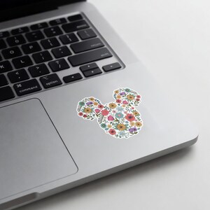 May include: A white sticker in the shape of Mickey Mouse's head, filled with colorful flowers. The flowers include red, yellow, pink, and blue blooms, creating a vibrant floral design. The sticker is placed on a silver laptop.