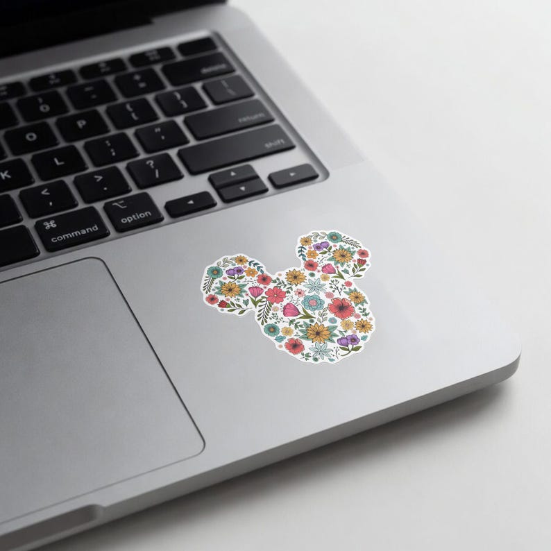 May include: A white sticker in the shape of Mickey Mouse's head, filled with colorful flowers. The flowers include red, yellow, pink, and blue blooms, creating a vibrant floral design. The sticker is placed on a silver laptop.