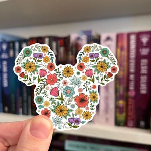 May include: A floral sticker in the shape of a mouse head. The sticker is filled with colorful flowers in shades of yellow, pink, orange, blue, and purple. The sticker is held in front of a bookshelf.