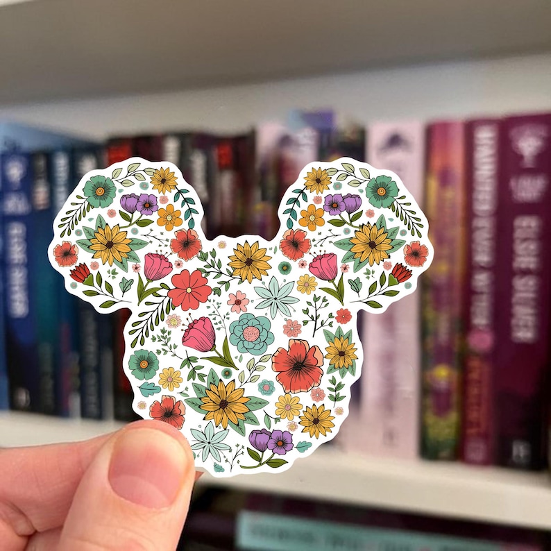 May include: A floral sticker in the shape of a mouse head. The sticker is filled with colorful flowers in shades of yellow, pink, orange, blue, and purple. The sticker is held in front of a bookshelf.