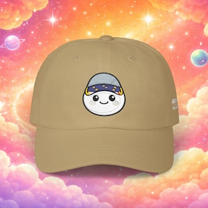 May include: A tan baseball cap featuring an embroidered cartoon character with a smiling face and a blue and gold head covering. The cap has a curved brim and a structured crown. The text "ART" is visible on the side.
