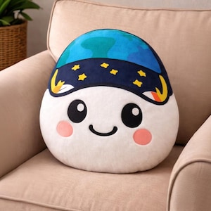 May include: A plush, egg-shaped pillow with a smiling face and a blue and navy blue design resembling a planet and starry night. The pillow has black eyes, pink cheeks, and a curved black mouth. It rests on a beige armchair.