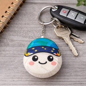 May include: A round keychain with a smiling face design. The keychain features a blue and yellow hat with stars, a white face, and pink cheeks. The keychain is attached to a silver chain and a keyring with keys and a car key fob.
