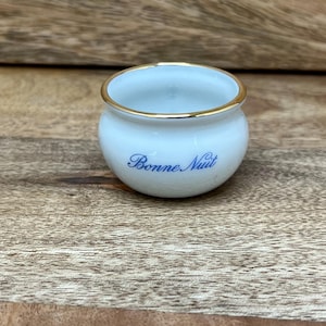 May include: Small, white ceramic bowl with a gold rim. The bowl has the words "Bonne Nuit" in blue script. The bowl is sitting on a wooden surface.