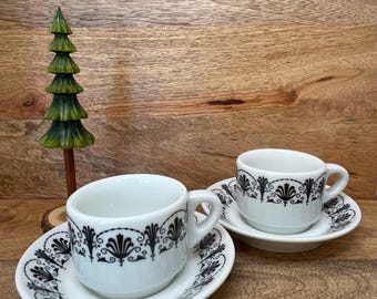May include: Two white ceramic espresso cups and matching saucers featuring a black decorative filigree pattern around the rims. The cups have a rounded handle and a smooth finish. A small green wooden tree ornament stands beside the cups.