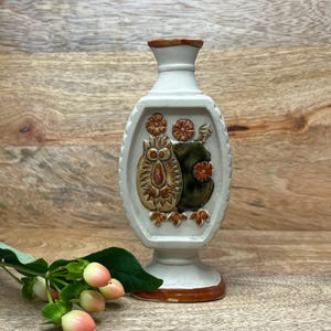 Può includere: Un vaso soliflore in ceramica color crema con collo stretto e base svasata. La parte anteriore presenta un rilievo in rilievo di un gufo stilizzato con accenti di smalto marrone e verde, circondato da piccoli motivi floreali arancioni.