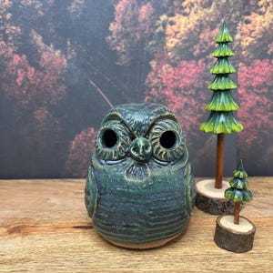 May include: A ceramic owl figurine in shades of green, with detailed facial features and large eye sockets. Beside it are two small wooden tree figurines, one tall and one short, with green painted branches. The background features a blurred forest scene.