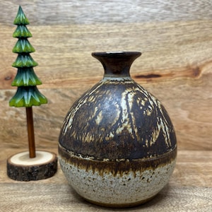May include: A round ceramic vase with a narrow neck, featuring a dark brown upper section with textured patterns and a speckled light beige lower section. Beside it stands a small, carved green wooden tree on a circular wood slice base.