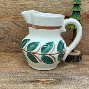 May include: A small, off-white ceramic pitcher with a brown band near the top and a green and brown leaf design. The pitcher has a curved handle and a spout for pouring. A small decorative green tree is in the background.