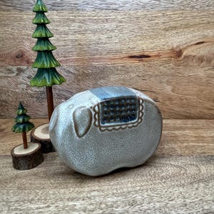 Może przedstawiać: Ceramiczna figurka słonia z nakrapianym jasnoszarym szkliwem, posiadająca ciemnoniebieskie prostokątne siodło z wzorem w kratkę. Obok stoją dwie rzeźbione drewniane sosny o różnych wysokościach, każda umieszczona na małej okrągłej drewnianej podstawie.