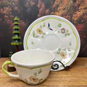 May include: A white teacup and saucer set with a floral design featuring yellow flowers and green leaves. The cup has a green handle and the saucer has a scalloped edge with a blue and green border. The set is on a wooden surface.
