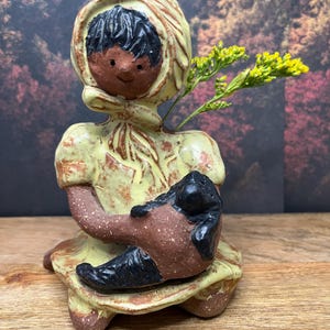 Może przedstawiać: Ceramiczna figurka osoby w kapturze trzymającej czarny przedmiot, prawdopodobnie lalkę lub zwierzę. Figurka jest głównie żółta i brązowa, z uśmiechniętą twarzą i czarnymi włosami. Żółte kwiaty umieszczono w figurce.