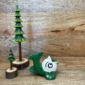 May include: A green and white ceramic dolphin figurine with a black eye and a small opening. Two wooden miniature Christmas trees with green painted details and a wooden base. The items are on a wooden surface.