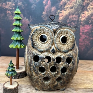 May include: A ceramic owl-shaped candle holder with a distressed brown and black finish. The owl has large eye openings and several circular cutouts. Two small wooden tree figurines are also present, one tall and one short, both painted green.