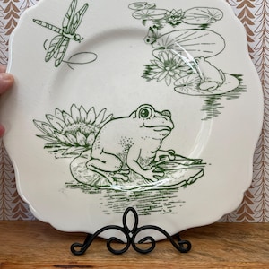Może przedstawiać: Biały ceramiczny talerz z falistą krawędzią, z zieloną ilustracją żaby na liściu lilii wodnej, ważki i lilii wodnych. Talerz jest prezentowany na czarnym metalowym stojaku, z drewnianą powierzchnią na pierwszym planie.