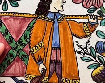Könnte beinhalten: Handbemalte Keramikplatte mit einer Figur in einem orangefarbenen Mantel und Hut, die einen Stab trägt. Die Figur ist von floralen und geometrischen Designs in Rot, Grün, Blau und Gelb umgeben. Die Platte hat einen blauen Rand und einen weißen Hintergrund mit Craquelé-Effekt.