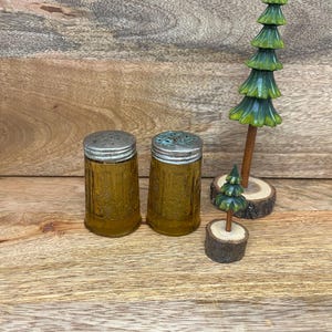 May include: Two amber-colored glass salt and pepper shakers with metal lids. A wooden decorative tree and a smaller tree on a wooden base are also present. The items are arranged on a wooden surface.