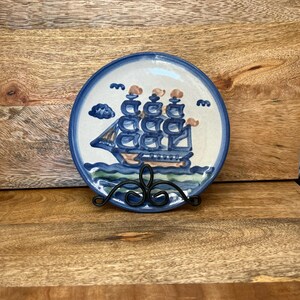 Puede incluir: Un plato redondo de cerámica pintado a mano con un diseño de velero azul y marrón. El plato tiene un borde azul y se exhibe en un soporte de metal negro. La obra de arte incluye nubes y olas.