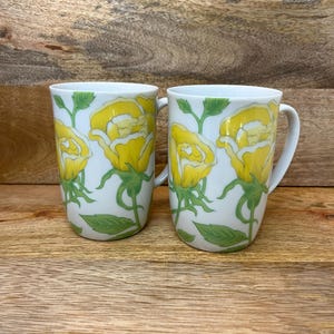 Puede incluir: Dos tazas de cerámica blanca con un diseño de rosas amarillas. Las rosas están detalladas con tallos y hojas verdes. Las tazas tienen una forma sencilla y elegante con un asa blanca.