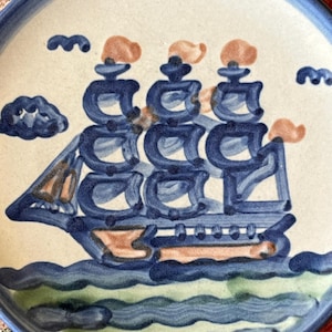 May include: A ceramic plate with a hand-painted nautical design. The plate features a blue and white color scheme with a ship sailing on blue waves. The ship has three masts with orange sails and is surrounded by blue clouds.
