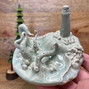 以下が含まれることがあります： ライトブルーの島にタコと灯台が描かれた陶器の彫刻。タコには複数の触手があり、灯台は円筒形です。彫刻は木製の表面にあります。