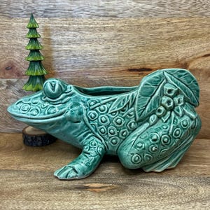 May include: A teal ceramic frog planter with detailed textures and a wide opening for plants. The frog has a detailed design and is placed on a wooden surface. A small green decorative tree is in the background.