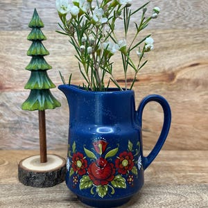 May include: A dark blue ceramic pitcher featuring a hand-painted floral motif with red roses and green leaves, containing white flowers. Beside the pitcher stands a small, carved wooden evergreen tree on a circular log base.