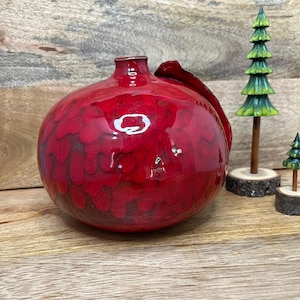 May include: A round, red ceramic vase with a glossy finish and a handle. The vase has a textured surface and is displayed with small decorative wooden Christmas trees.