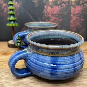 Può includere: Una tazza di ceramica bianca con manico nero e la scritta "But First, Coffee" in grassetto nero. La tazza è piena di un liquido scuro, probabilmente caffè, e poggia su una superficie chiara.