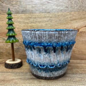 Peut inclure: Un pot en céramique au corps conique texturé, doté d'un vernis dégoulinant blanc et bleu, marqué par des stries verticales et des accents bleu foncé. À côté se trouve un petit sapin en bois sculpté sur un socle rond.