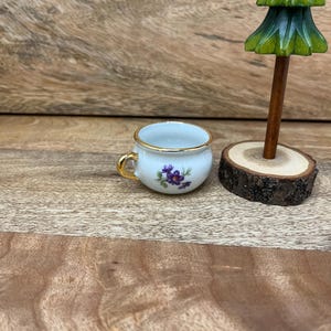May include: A small, white teacup with a gold rim and handle, adorned with a purple floral design. Beside it, a miniature wooden Christmas tree stands on a wood slice base, set against a wooden background.