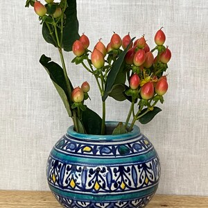 May include: A round ceramic vase featuring a hand-painted pattern of dark blue, teal, and yellow geometric shapes and floral motifs. The vase contains several stems of Hypericum berries with small, round, coral-pink fruits and green leaves.