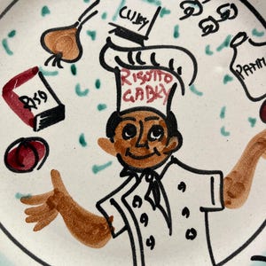 May include: A white ceramic plate with a cartoon chef illustration. The chef wears a white uniform and hat with "Risotto Gaby" on it. The plate has drawings of ingredients like onions, tomatoes, and spices, with the word "Curry" visible.