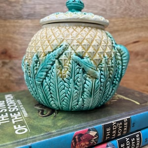 Può includere: Un barattolo in ceramica rotondo con coperchio, caratterizzato da una parte superiore color crema con texture ad ananas e una base verde brillante decorata con motivi a foglia di felce in rilievo. Il coperchio ha un piccolo pomello rotondo verde acqua.