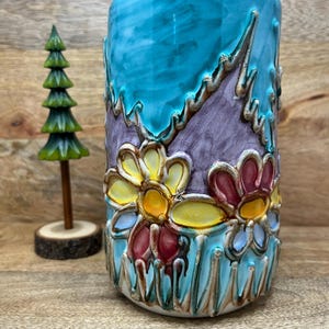May include: A cylindrical ceramic vase featuring a vibrant turquoise blue background with raised, hand-painted floral motifs in yellow, red, and purple. The design includes textured, relief-style outlines and a small, separate miniature green pine tree.