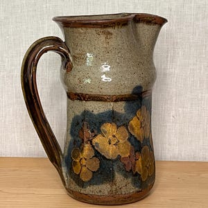 Può includere: Una brocca in gres con smalto beige maculato, manico e bordo marrone scuro. La parte inferiore presenta un motivo floreale dipinto a mano nei toni del giallo senape e del blu profondo su una base strutturata di colore grigio-marrone.