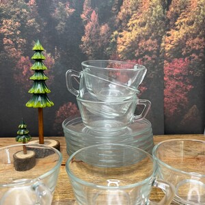 May include: A stack of clear glass teacups and saucers. The teacups have a rounded shape with a handle. The saucers are flat and round. A small, green and yellow decorative tree is in the background.